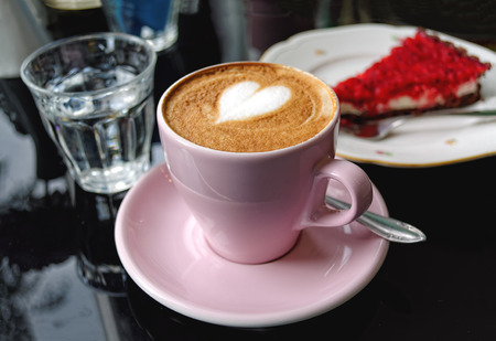Pink cup of cappuccino, with milk foam in form of heart, on the table in the cafe with cheesecake in the background.の写真素材