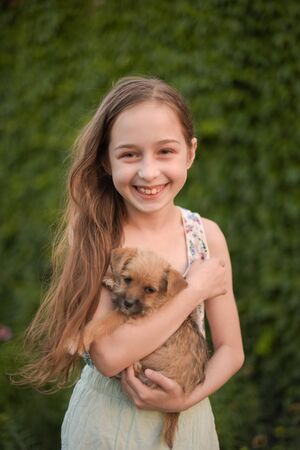 A little blond girl with her pet dog outdooors in park. The girl loves a little puppy. A dog in the arms of a child. Summer rest.の写真素材