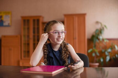 Schoolgirl sitting at the table with textbooks in the classroom. Girl in a blue dress with pigtails. A little girl listens to music on headphones from a phone. Modern children. 4th grade schoolの写真素材
