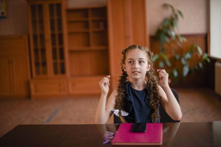 Schoolgirl sitting at the table with textbooks in the classroom. Girl in a blue dress with pigtails. A little girl listens to music on headphones from a phone. Modern children. 4th grade schoolの写真素材