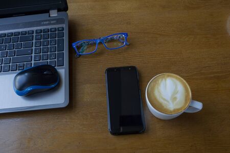 Workspace with laptop, a cup of coffee and a mouse for a laptop, phone, glasses on a white wooden table. Gray laptop, blue mouse for working at a computer, blue glasses for vision, cappuccino.の写真素材