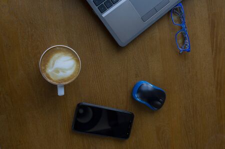 Workspace with laptop, a cup of coffee and a mouse for a laptop, phone, glasses on a white wooden table. Gray laptop, blue mouse for working at a computer, blue glasses for vision, cappuccino.の写真素材