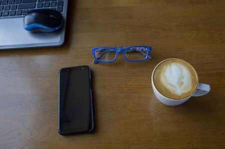 Workspace with laptop, a cup of coffee and a mouse for a laptop, phone, glasses on a white wooden table. Gray laptop, blue mouse for working at a computer, blue glasses for vision, cappuccino.の写真素材