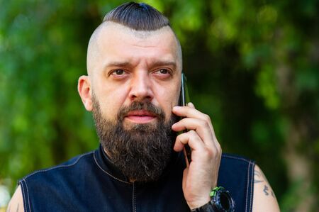 Man talking on mobile phone over green background. A man in dark clothes with a fashionable haircut and beard is talking on a black mobile phone.の写真素材