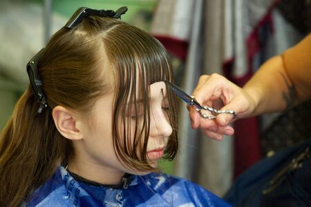 Hairdresser making a hair style to cute little girl. The girl cuts off the bangs. Work hairdresser. Change the image. Change hairstyle. Fashionable baby. A series of photos of haircuts baby bangs.の写真素材