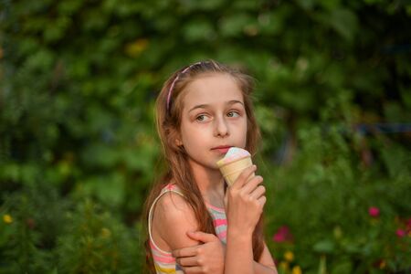 A little girl of 9 years old in a bright striped dress is eating a bright ice cream at an outdoor in a colorful striped bright dress. Sunny summer hot weather. The little blonde laughs wholeheartedly.の写真素材