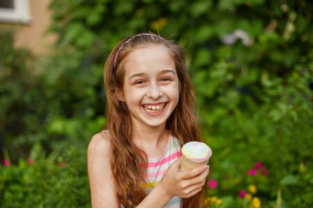 A little girl of 9 years old in a bright striped dress is eating a bright ice cream at an outdoor in a colorful striped bright dress. Sunny summer hot weather. The little blonde laughs wholeheartedly.の写真素材
