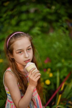 A little girl of 9 years old in a bright striped dress is eating a bright ice cream at an outdoor in a colorful striped bright dress. Sunny summer hot weather. The little blonde laughs wholeheartedly.の写真素材