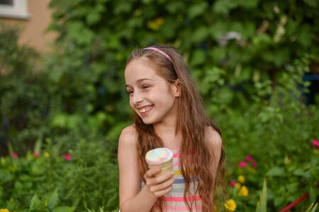 A little girl of 9 years old in a bright striped dress is eating a bright ice cream at an outdoor in a colorful striped bright dress. Sunny summer hot weather. The little blonde laughs wholeheartedly.の写真素材