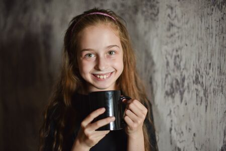 Little girl holding a coffee cup in black t-shirt. Mock up, perfect for putting your design on. Beautiful girl with long blond hair 9 years old drinks a drink from a black cup. A cute baby. teensの写真素材