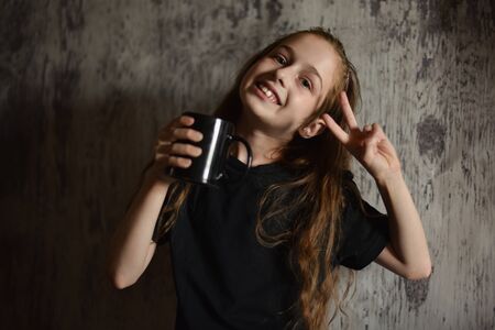 Little girl holding a coffee cup in black t-shirt. Mock up, perfect for putting your design on. Beautiful girl with long blond hair 9 years old drinks a drink from a black cup. A cute baby. teensの写真素材