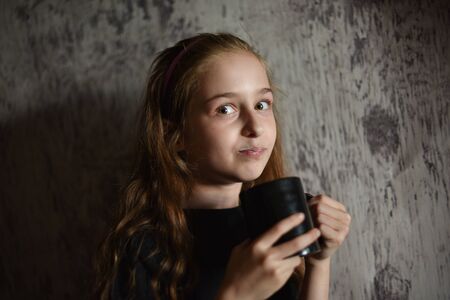 Little girl holding a coffee cup in black t-shirt. Mock up, perfect for putting your design on. Beautiful girl with long blond hair 9 years old drinks a drink from a black cup. A cute baby. teensの写真素材