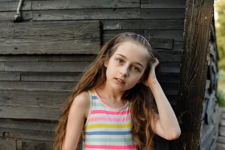 Pensive little child with long hair resting on wooden deck, looking aside while dreaming about something. Cute baby in a white dress in a multicolored stripes. Beautiful girl on the wooden background.の写真素材