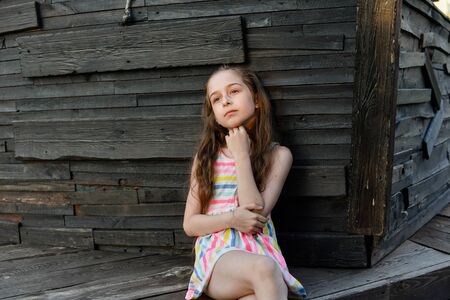 Pensive little child with long hair resting on wooden deck, looking aside while dreaming about something. Cute baby in a white dress in a multicolored stripes. Beautiful girl on the wooden background.の写真素材