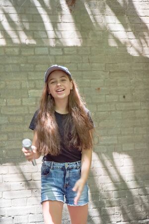 Beautiful little girl with microphone on brick wall background.The girl in denim shorts sings into a microphone on a city street. Beautiful brunette. Vote. Long hair in a child. Lifestyleの写真素材