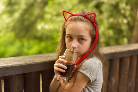 The girl wore a hoop cat ears and a red strand. A child walks in a cafe and drinks a drink from a glass. 9-year-old girl Modern children. Summer terrace. sunny day. Modern children.の写真素材