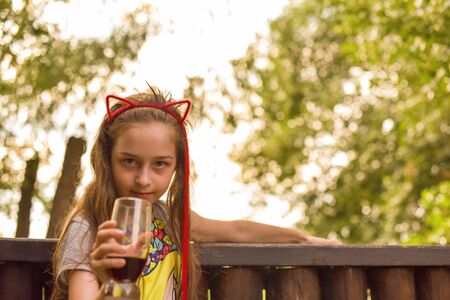 The girl wore a hoop cat ears and a red strand. A child walks in a cafe and drinks a drink from a glass. 9-year-old girl Modern children. Summer terrace. sunny day. Modern children.の写真素材