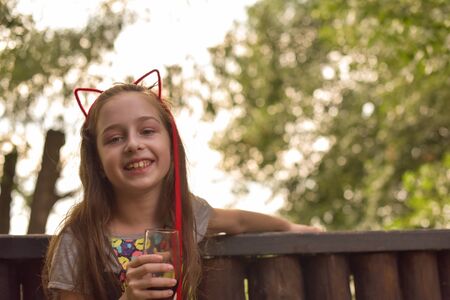 The girl wore a hoop cat ears and a red strand. A child walks in a cafe and drinks a drink from a glass. 9-year-old girl Modern children. Summer terrace. sunny day. Modern children.の写真素材