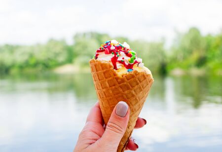 funny creative concept of wafer cone with ice cream covered and strewed sprinkles. Ice cream horn on the background of the river. Ice cream horn in nature in a woman's hand with french manicureの写真素材