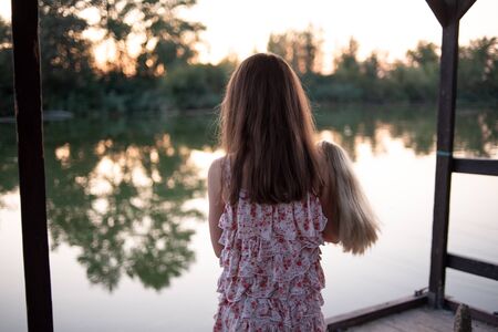 Girl in dress walking on the promenade.Little girl with a doll by the river.A girl of 9 years old is standing by the river on a wooden deck.Relax Rest. Girl in the summer in a dress.Beautiful hairの写真素材