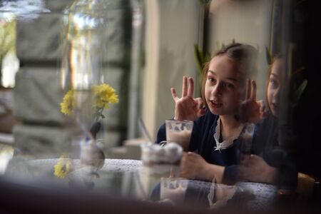 Little girl drinking hot chocolate in a cafe. schoolgirl drinks cocoa in a cafe. girl after classes in a coffee house drinks cocoa and eats a dessert. A girl is sitting by the window. Back to schoolの写真素材