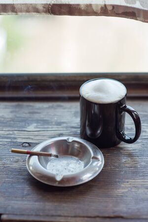 coffee and burning cigarette in the photo. Cup of cappuccino and cigarette. Black cup with latte. Coffee with milk foam stands on a wooden tabletop. Brown metal cigar ashtray.Coffee milk foam. cigarの写真素材