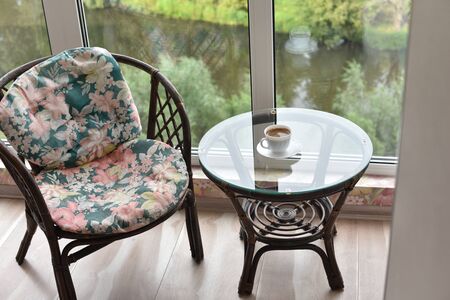Cup of coffee on a glass table by the window and an armchair.Table, chair, cups and ashtray on...
