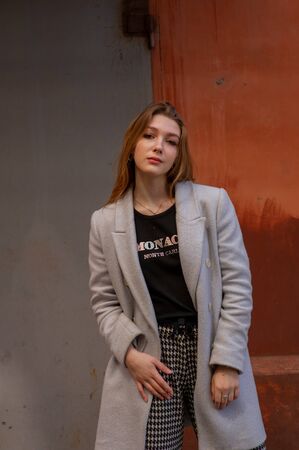 Stylish young pretty girl in a gray coat looks at the camera and smiles slightly against the...