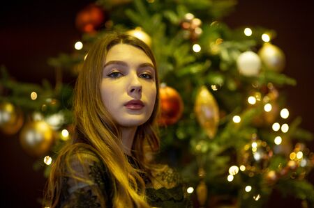 New Year's mood in a blonde girl. pretty caucasian young girl, seductive lipstick and wavy hair standing in cafe. Beautiful cute woman and christmas tree. Girl in evening black dress by the Christmasの写真素材