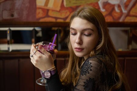 Girl blonde in black drinks a cocktail in a cafe. Girl in a black dress with a cocktail in a cafe. A...