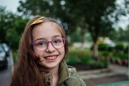 Little girl on the street against a background of green foliage.Girl with long hair. Child 9-10 years old girl.Girl 9 years old in the spring. A teenager in the spring on a walk. Military style jacketの写真素材