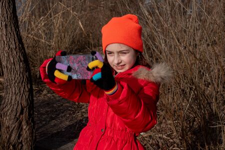 Girl dressed in red jacket, poses for making selfie or photo of herself with smartphone. People and...