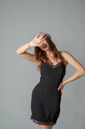 Glamorous young woman wears fashionable dress posing with fluttering hair at grey wall. Girl put forward hand in a black dress and a gray cap. Beautiful young girl with brown curly hairs. portraitの写真素材