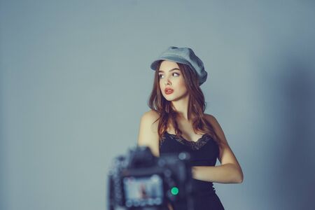 Glamorous young woman wears fashionable dress posing with fluttering hair at grey wall.Girl put forward hand in a black dress and a gray cap.A girl is photographed on a camera or I am shooting a videoの写真素材