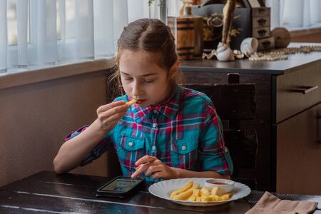 girl eating french-fries. Girl eating french fries in a cafe. A girl of 9 years old has lunch with french fries. French fries, sauce, lunch, food. A schoolgirl is eating and looking at a smartphone.の写真素材