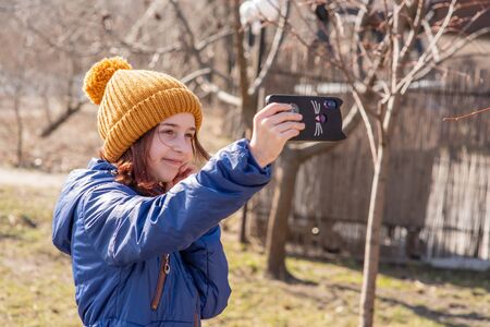 Girl dressed in jacket, poses for making selfie or photo of herself with smartphone. People and leisure concept. Girl makes selfie in a red jacket.Girl 9 years old with a smartphone in her hands. Teenの写真素材