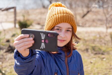Girl dressed in jacket, poses for making selfie or photo of herself with smartphone. People and leisure concept. Girl makes selfie in a red jacket.Girl 9 years old with a smartphone in her hands. Teenの写真素材