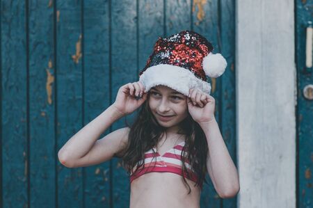 A child is playing with a santa claus hat. Happy little girl in Santa Claus hat outdoors in summer.Concept New Year or Christmas series of photos.Little girl in a Santa Claus hat by the poolの写真素材