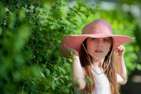 Cute teenage girl 8-9 years old. Teenager. Portrait of a little girl. Girl in a white dress and a hat on a background of green foliage.の写真素材