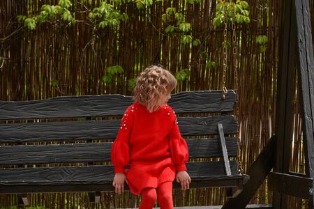 Girl 7 years old in red clothes on the street on a swing. Little curly blonde girl on a swing in spring. Schoolgirl. 1st grade student. Spring, nature, childhoodの写真素材