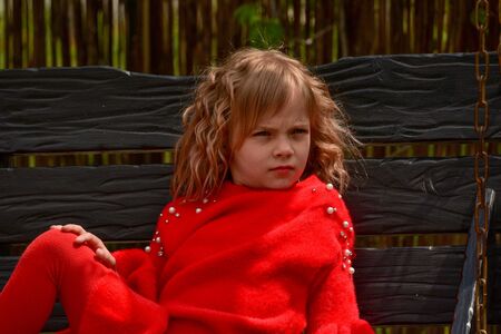 Girl 7 years old in red clothes on the street on a swing. Little curly blonde girl on a swing in spring. Schoolgirl. 1st grade student. Spring, nature, childhoodの写真素材