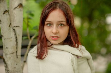 Portrait of a cute little girl nine years old. A little girl in a white coat stands by the birch. A...