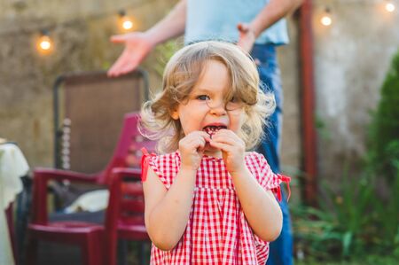Girl Eating Sausage At Family Barbeque. 2 years old girl eats sausage. Little girl on nature mouth a piece of sausage. A child on a walk. Portet baby girl 2 years old.の写真素材