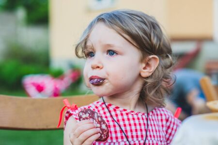 Girl Eating Sausage At Family Barbeque. 2 years old girl eats sausage. Little girl on nature mouth a piece of sausage. A child on a walk. Portet baby girl 2 years old.の写真素材