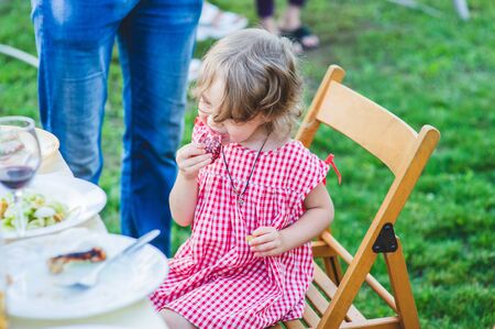Girl Eating Sausage At Family Barbeque. 2 years old girl eats sausage. Little girl on nature mouth a piece of sausage. A child on a walk. Portet baby girl 2 years old.の写真素材