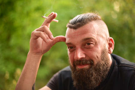 Man with a beard on a background of greenery. Large facial portrait of a bearded man of 40 years. Portrait full face. Man in a black T-shirt. A man smokes a cigarette.の写真素材