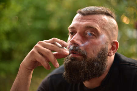 Man with a beard on a background of greenery. Large facial portrait of a bearded man of 40 years. Portrait full face. Man in a black T-shirt. A man smokes a cigarette.の写真素材