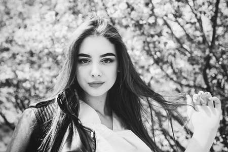 Young beautiful girl with long hair enjoys the beauty of spring nature near the blossoming sakura tree. Girl in a leather jacket in sakura. Black and white photo of a girl. Portraitの写真素材