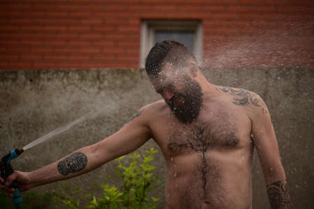 A man douses himself with water from a hose in the street. A man with a beard is watering. A guy in denim shorts. Summer, heat, waterの写真素材