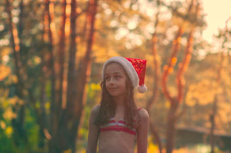 A child is playing with a santa claus hat. Happy little girl in Santa Claus hat outdoors in summer.Concept New Year or Christmas series of photos.Little girl in a Santa Claus hat by the poolの写真素材